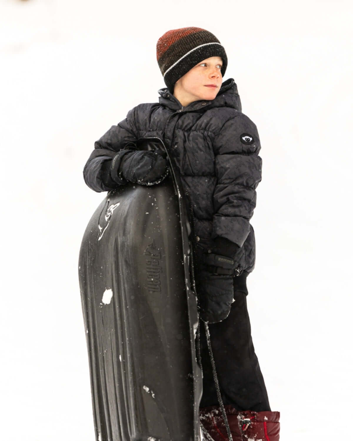 Stylish kid in a cozy puffy coat and hat, ready for winter fun with a sled in snowy outdoor adventure.