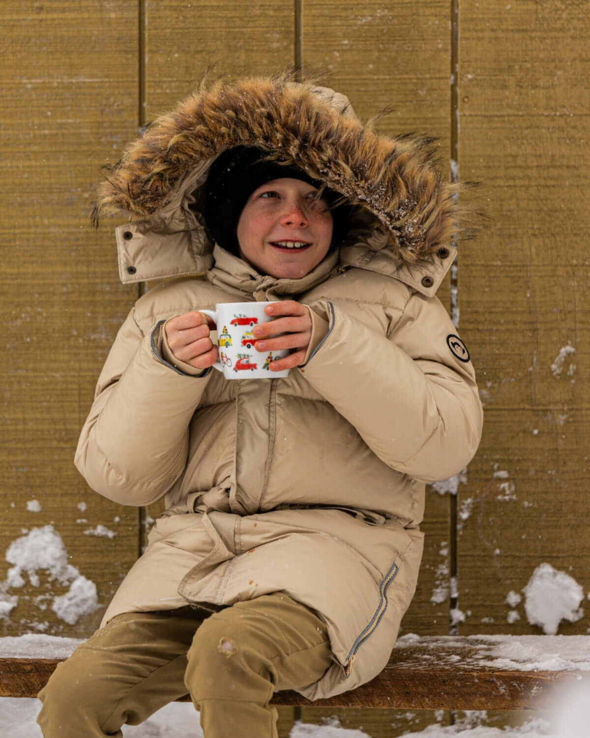 Cozy kid in an ivory puffer coat with faux fur hood, sipping hot cocoa in the snow, ready for winter adventures.