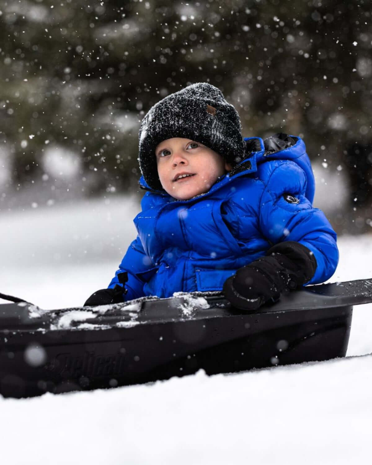 Joyful boy in a Reflex Blue Summit Puffer Coat sledding in the snow, showcasing stylish winter kids clothes.