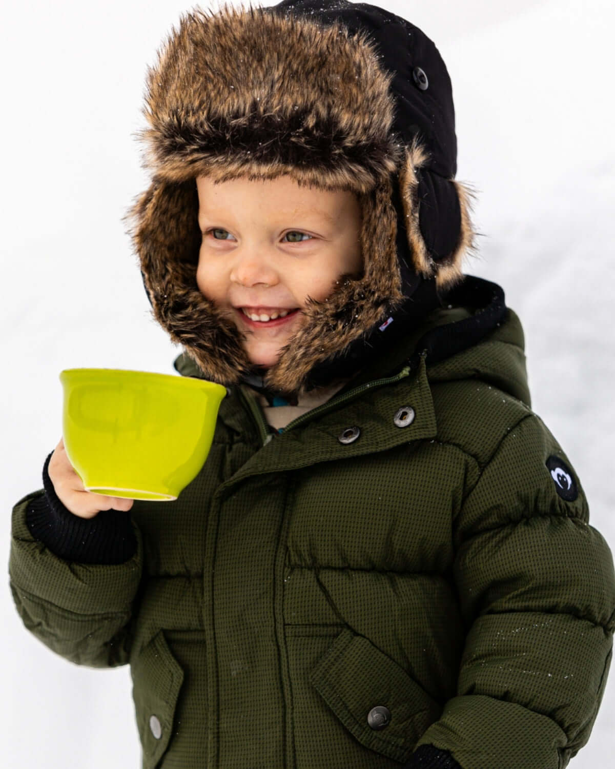 Happy kid in a Thunderstorm Puffy Coat, enjoying winter with a cozy hat and a bright green bowl.
