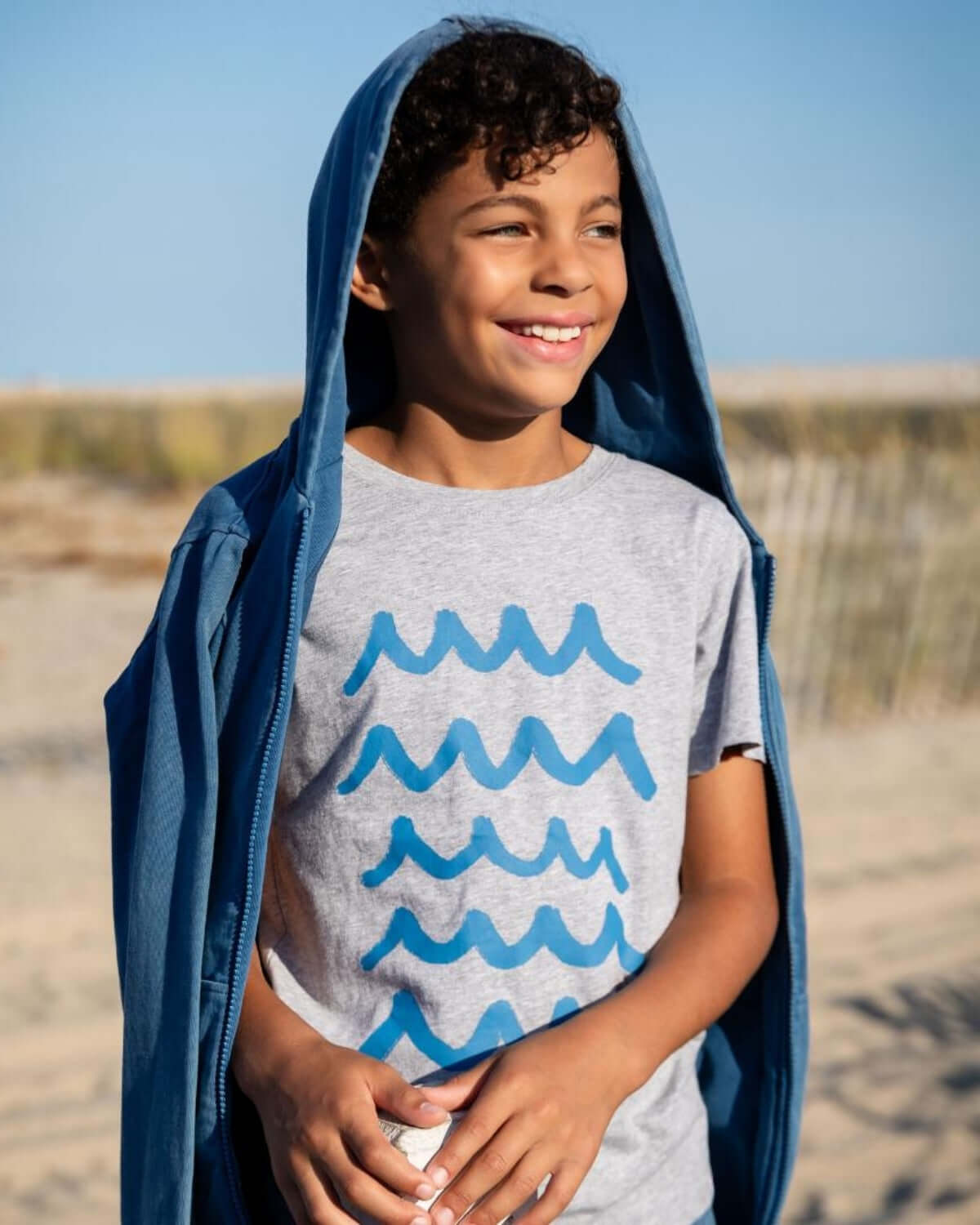 Happy boy in a sunwashed blue hoodie and wave-graphic tee, ready for beach fun in stylish kids' clothes.
