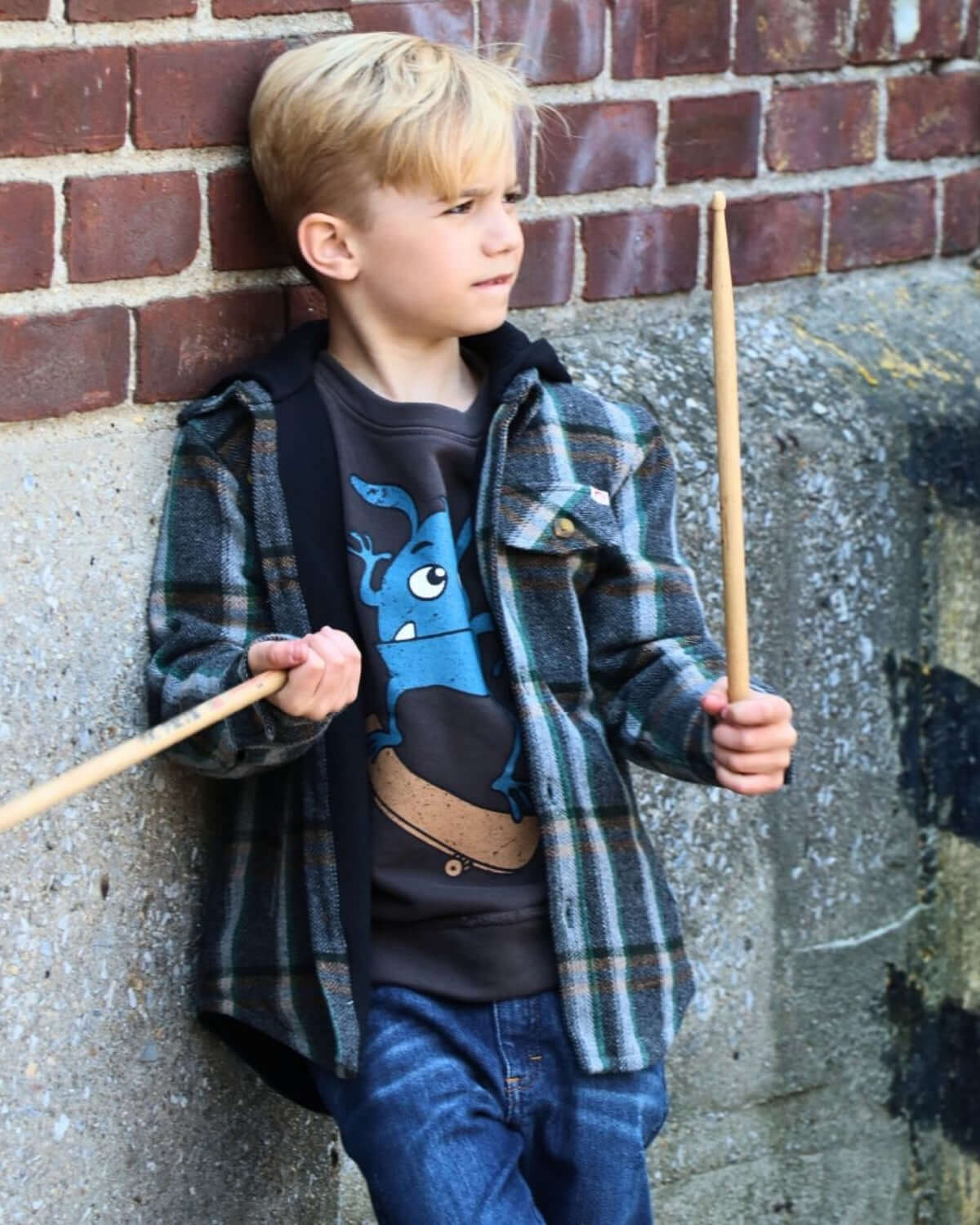 Boy wearing Woodland Glen Hooded Flannel, posing with drumsticks in a stylish outdoor setting against a brick wall.