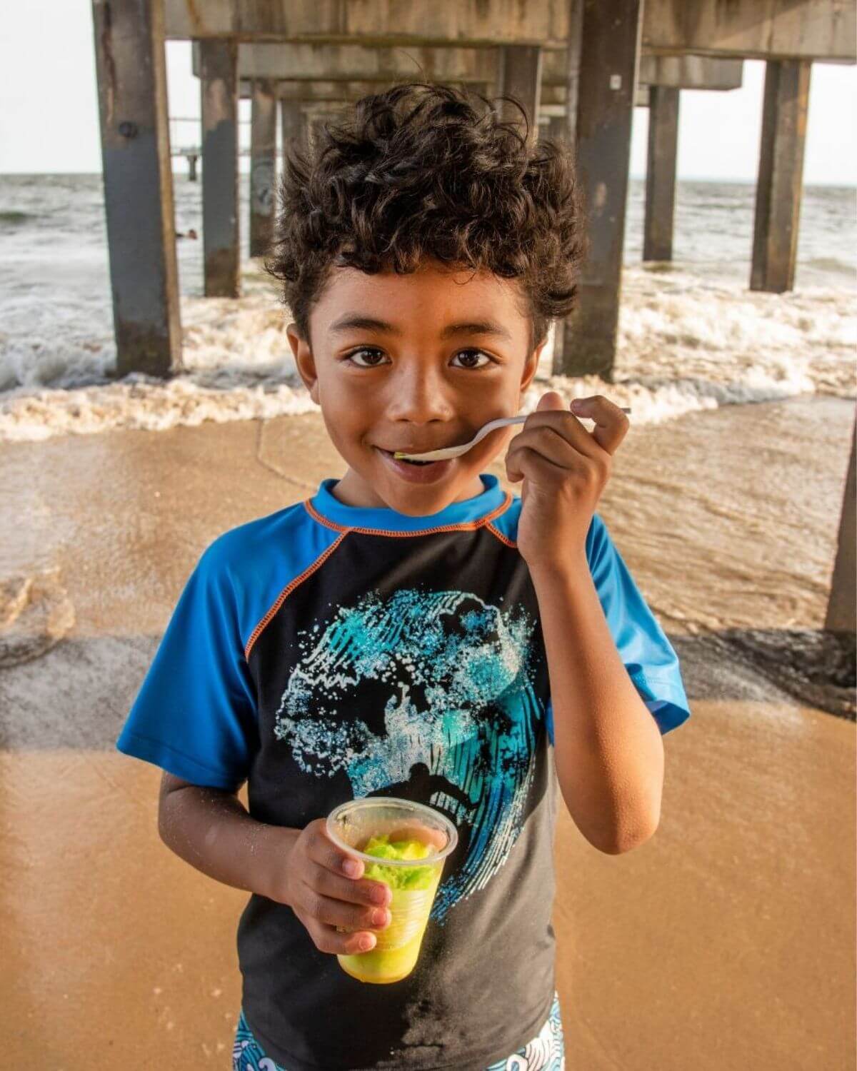 Child wearing Surfer Wave Blue Rash Guard enjoying a snack on the beach under a pier.
