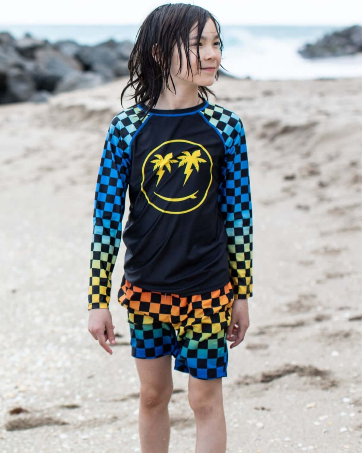 Cool kid sporting vibrant checkerboard swim trunks and a smiley palm tree rash guard at the beach—fun summer vibes!