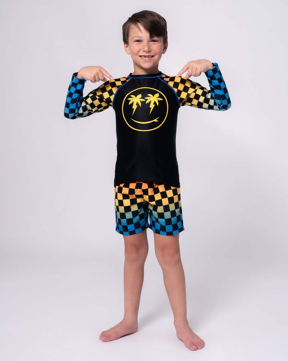 Stylish boy in vibrant checkerboard swim trunks and sun-soaked rash guard, ready for summer fun in high-quality kids clothes.