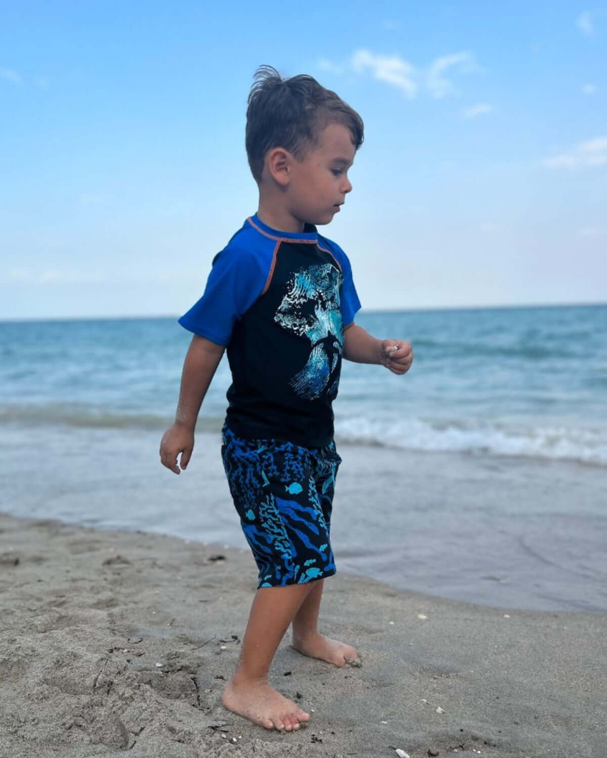 Boy wearing Coral Swim Trunks and rash guard playing on the beach by the water.