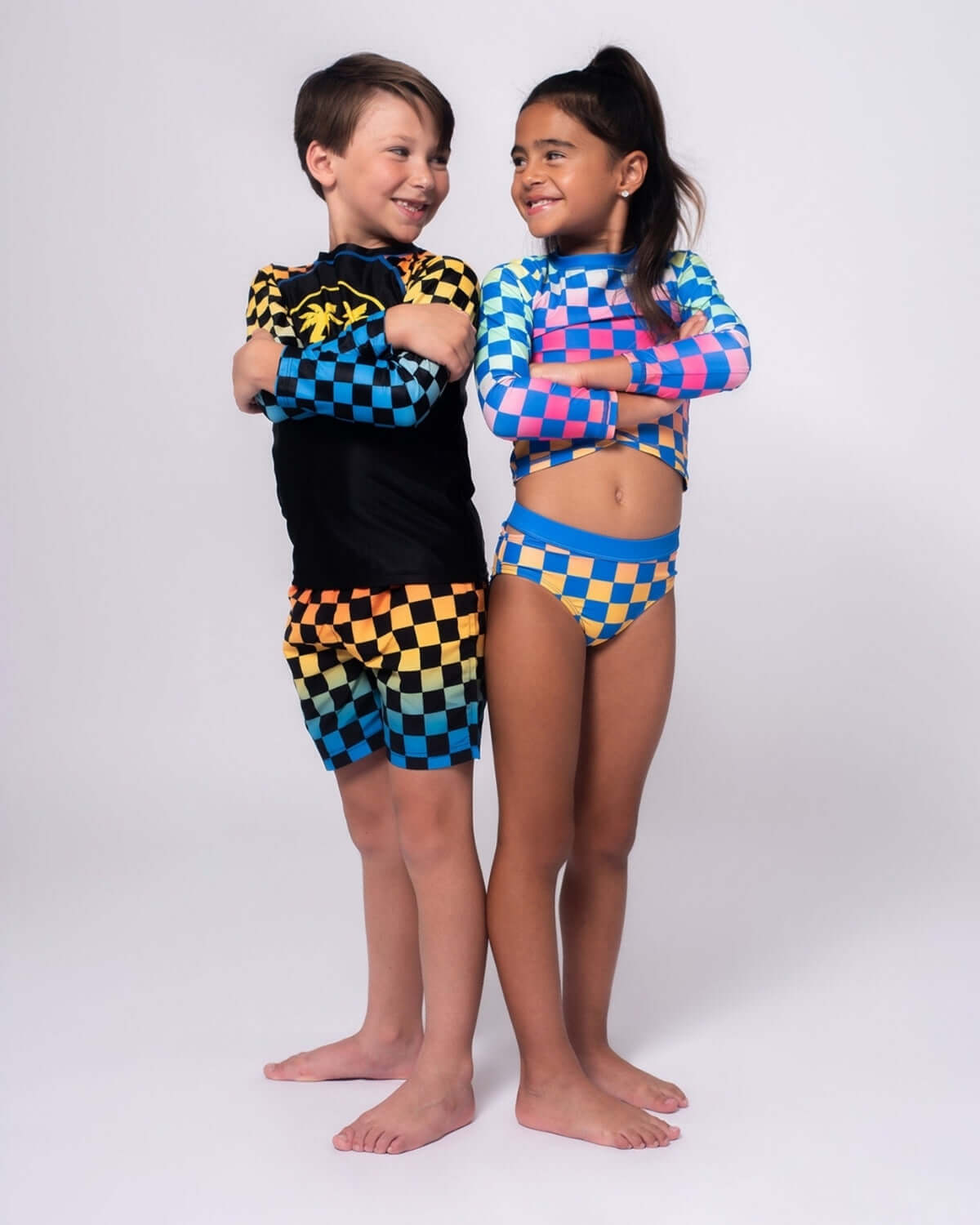 Kids wearing Happy Surfing Rash Guard and swimwear with checkerboard patterns, ready for fun in the sun.