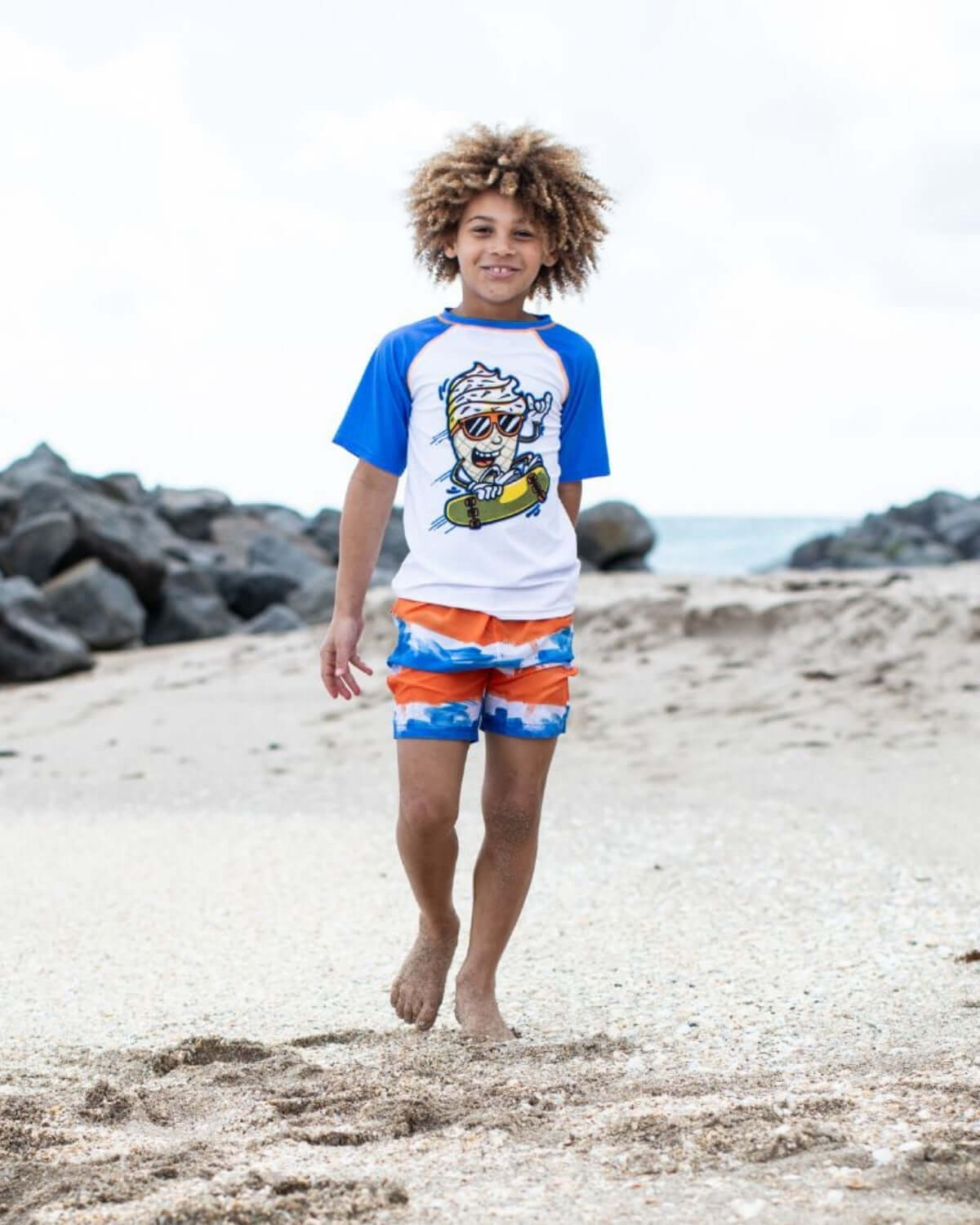 Boy wearing Soft Served Rash Guard and colorful swim trunks on the beach, enjoying summer fun.