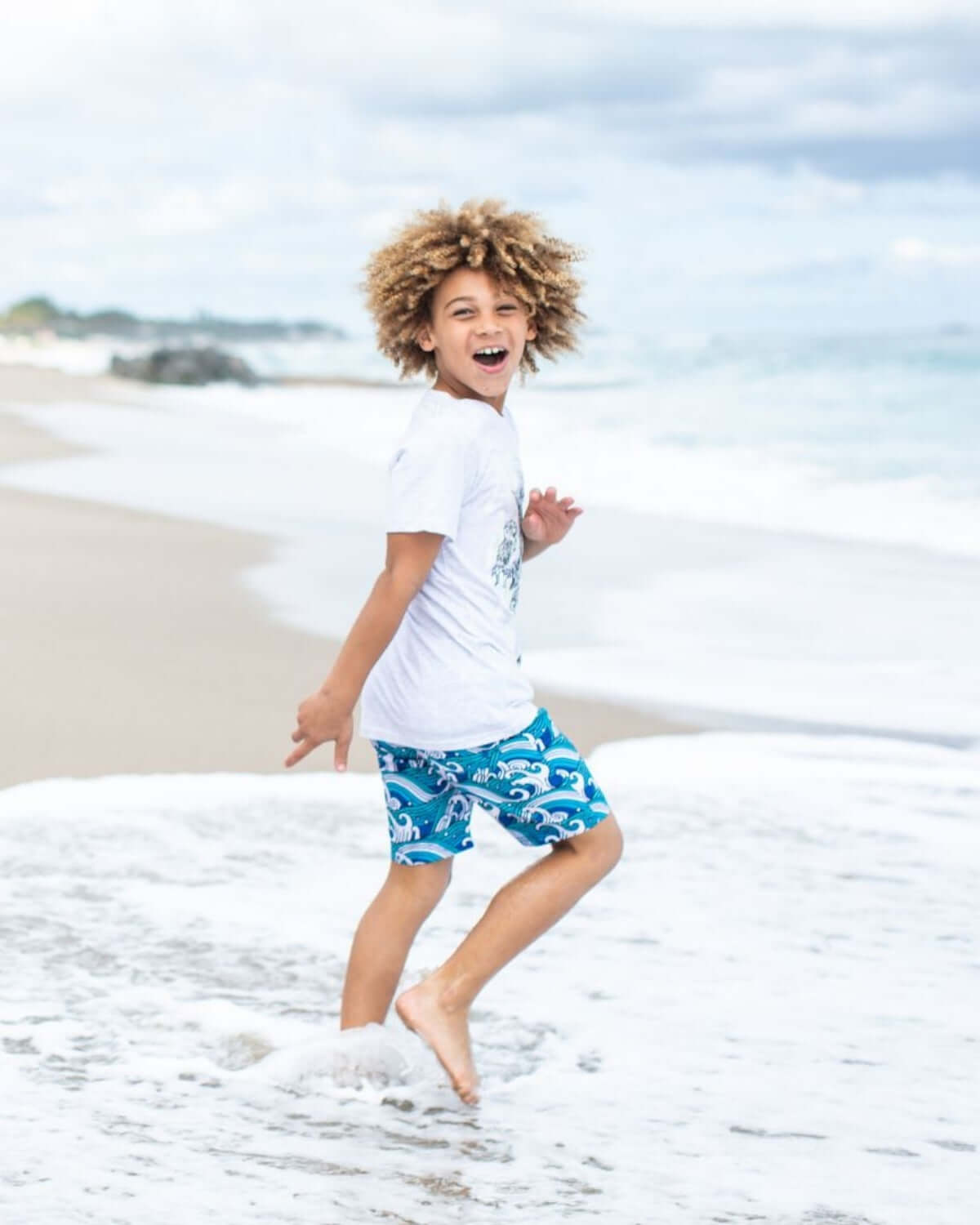 Boy running on the beach wearing Waves Swim Trunks and a white t-shirt, enjoying the summer sun.