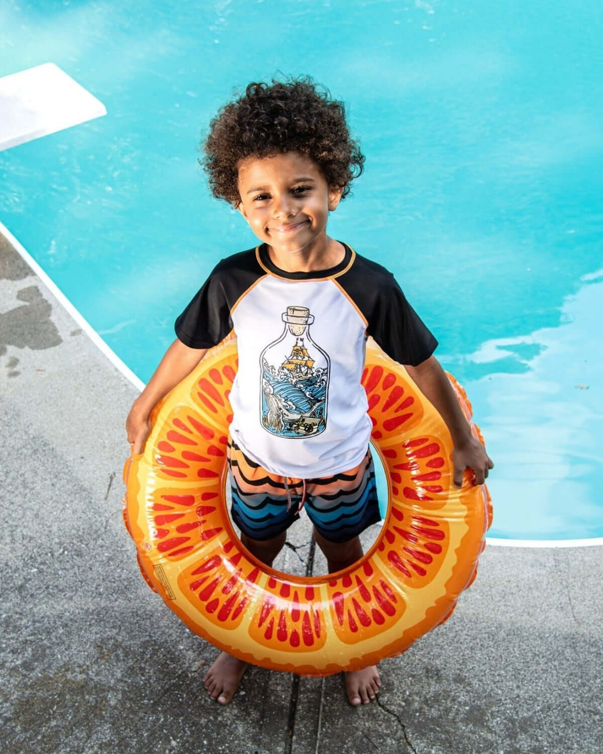 Joyful kid in colorful swim trunks and playful floatie, ready for fun by the pool—stylish kids clothes for summer adventures.