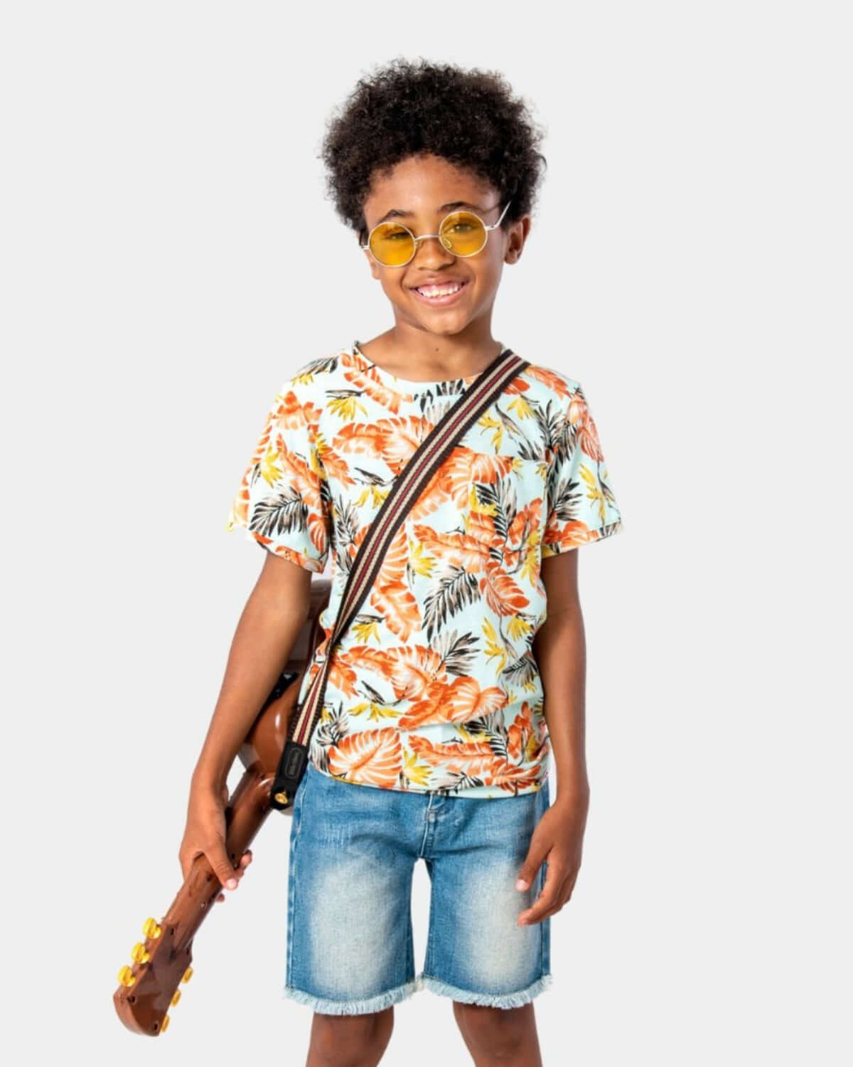 A child wearing a Tropics Concert Tee with a floral design, holding a ukulele and wearing yellow sunglasses.