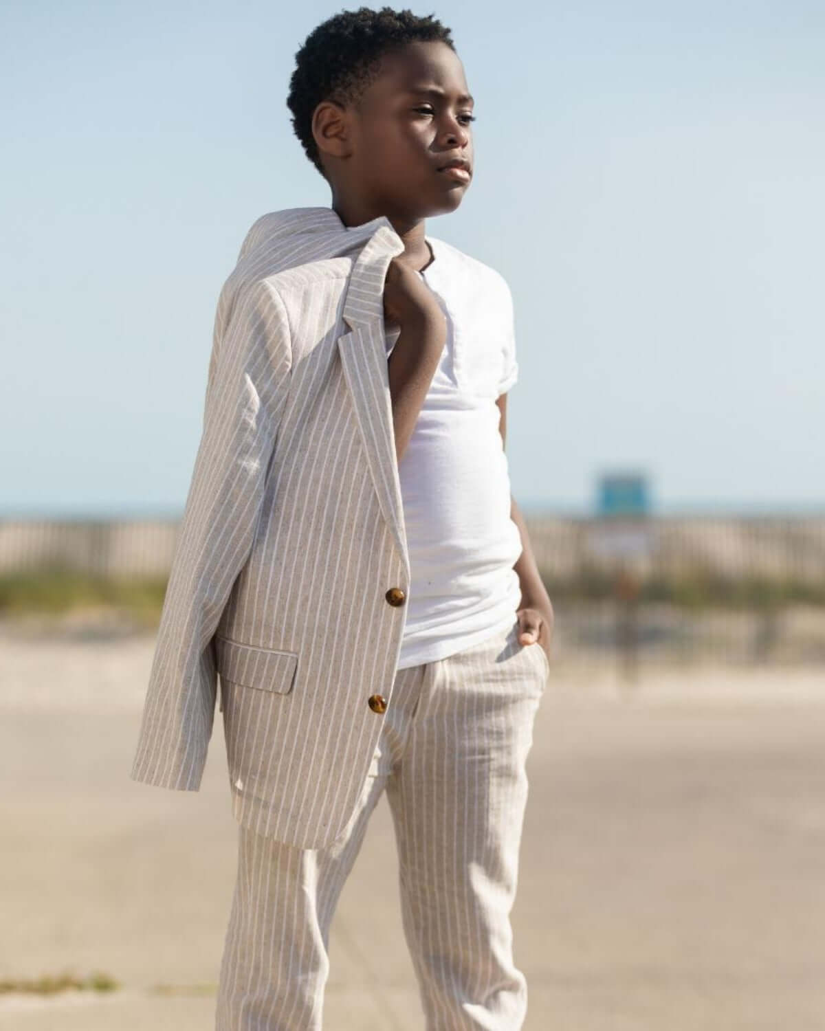Stylish young boy in a striped suit jacket and t-shirt, exuding confidence and charm by the beach.