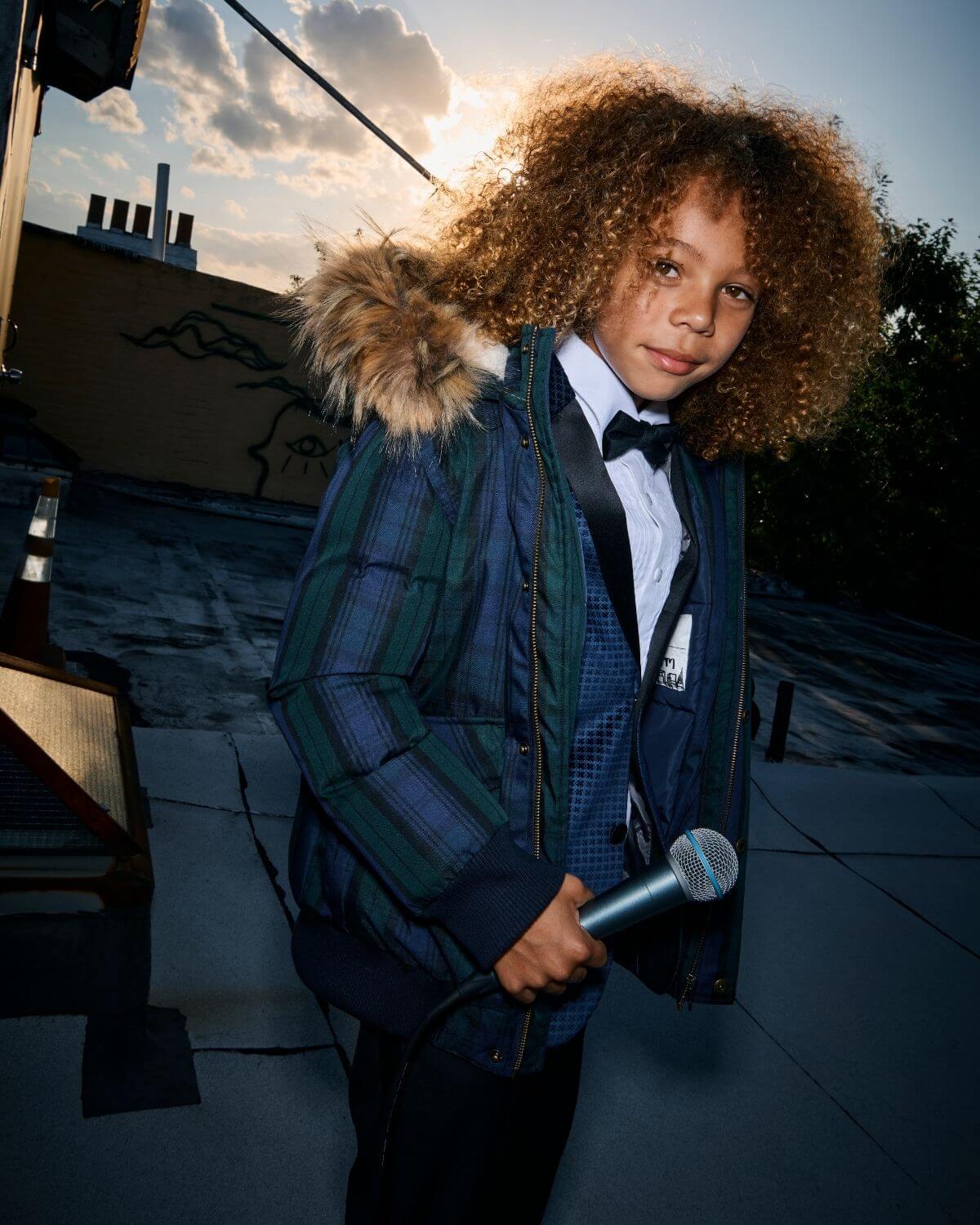 Stylish kid in a Blackwatch Tartan coat, ready for winter adventures with a microphone in hand, shining in the sunset.