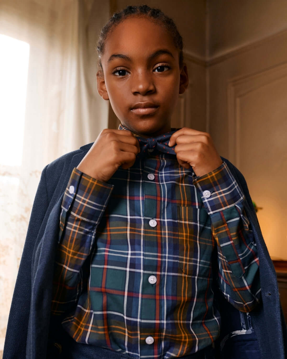 Child adjusting a Navy Darts Bow Tie, wearing a stylish plaid shirt and blazer for a dapper look.