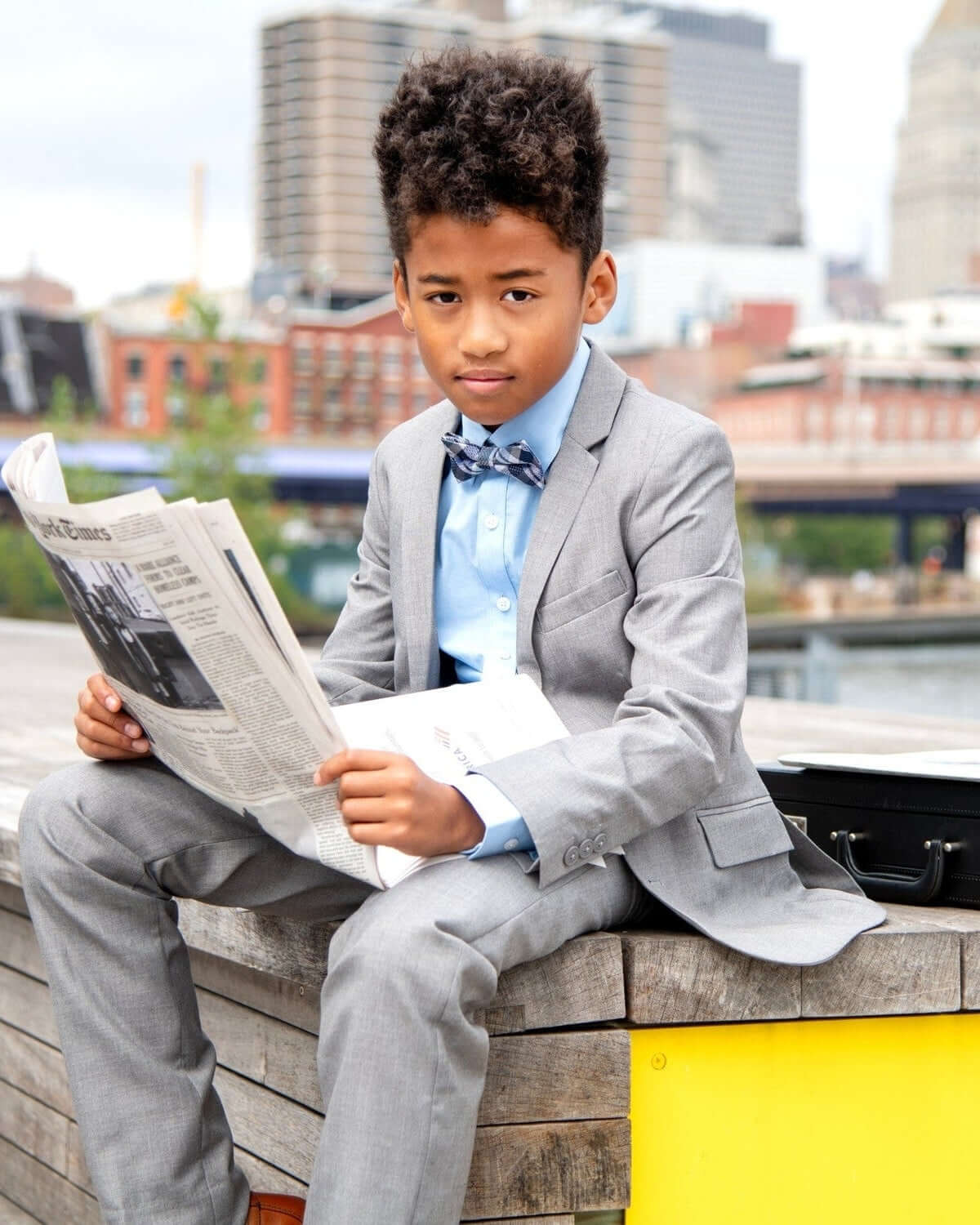 Stylish kid in a gray suit and navy/green gingham silk bow tie reading the newspaper by the waterfront.