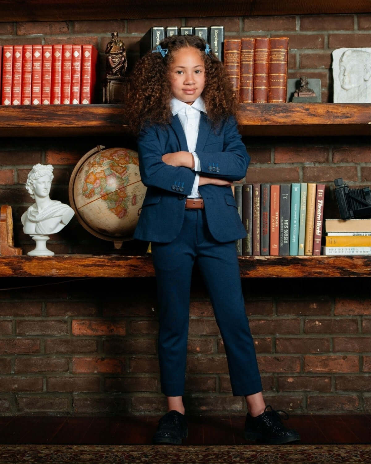 A stylish child wearing Blueprint Stretchy Suit Pants and a blazer, posing in front of a bookshelf.