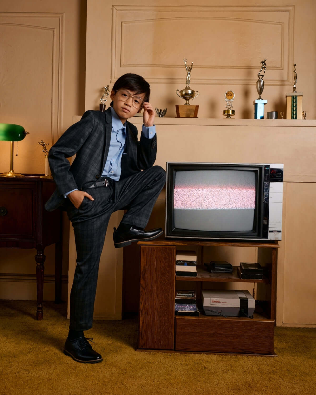Stylish kid in plaid suit pants strikes a confident pose by a vintage TV, showcasing modern charm and playful confidence.