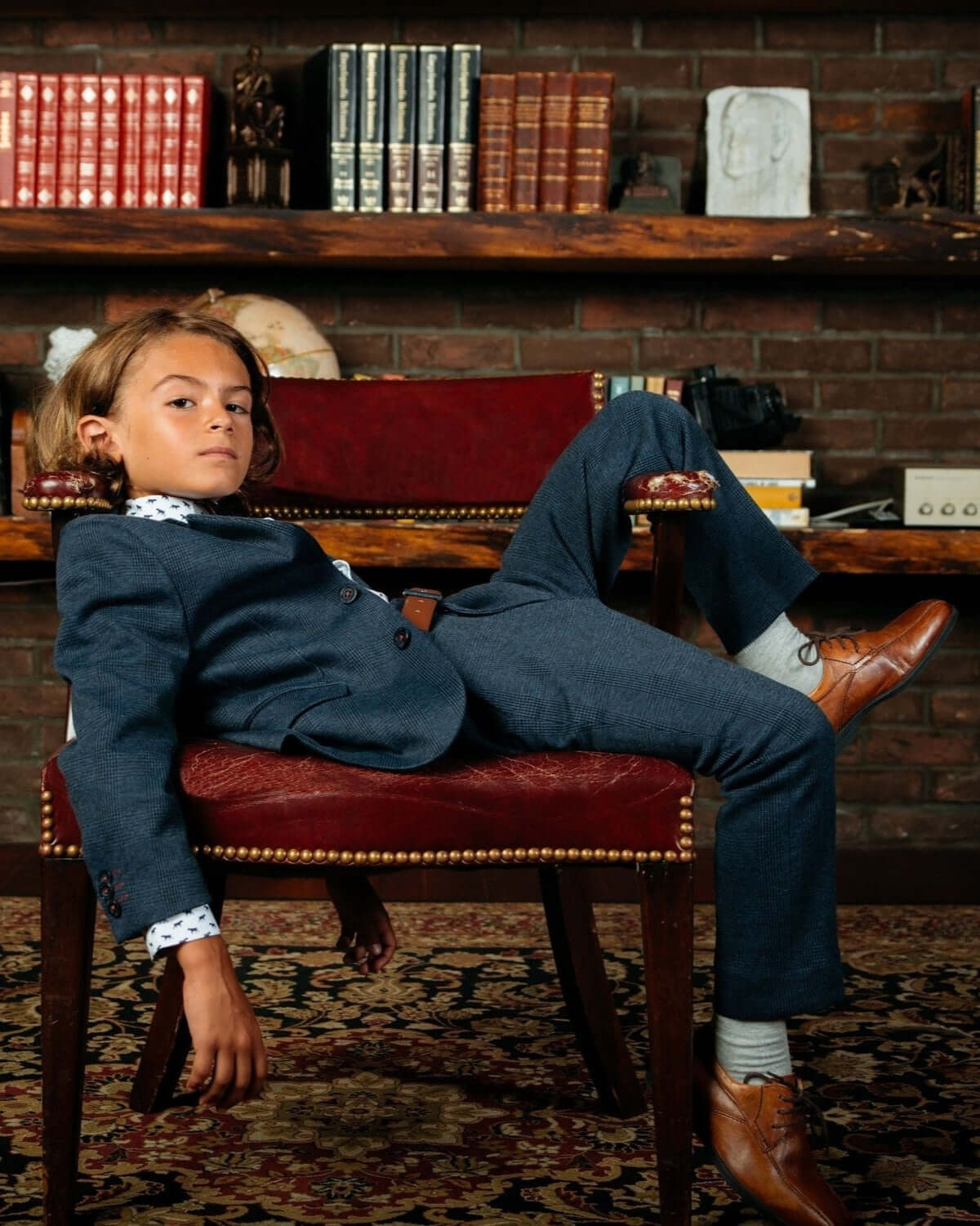 Stylish boy in Indigo Wales Check Stretchy Suit Pants sitting on a chair with a bookshelf in the background.