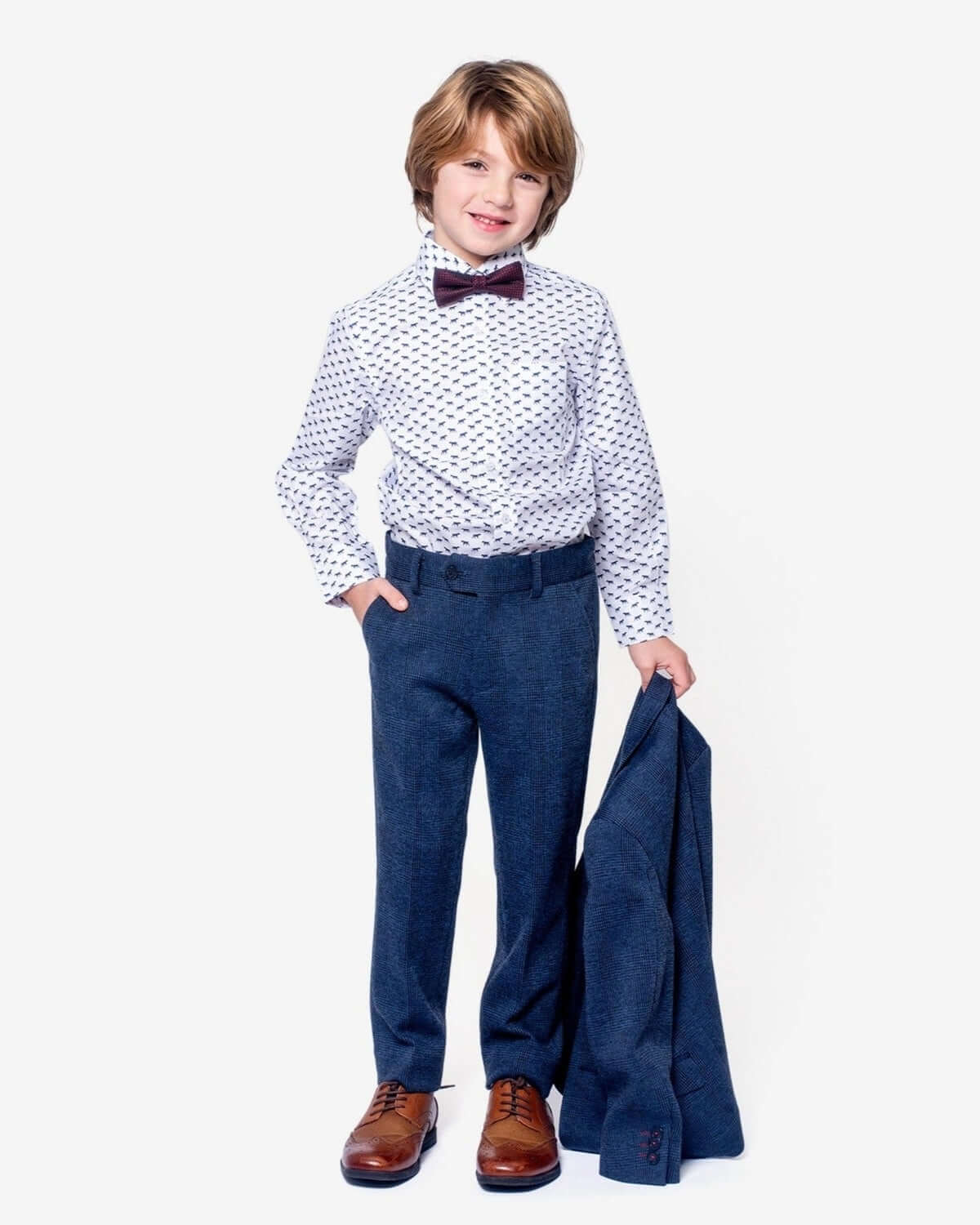 A boy wearing Indigo Wales Check Stretchy Suit Pants with a dress shirt and bow tie, holding a suit jacket.