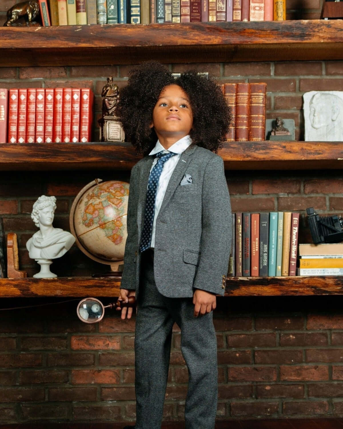 Stylish boy in a gray suit with a magnifying glass, posing confidently against a charming bookshelf backdrop.