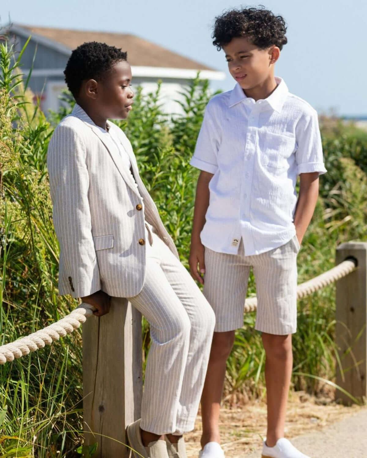 Two stylish boys in beach chic outfits: one in a linen suit, the other in a crisp button-up and shorts, soaking up summer vibes.