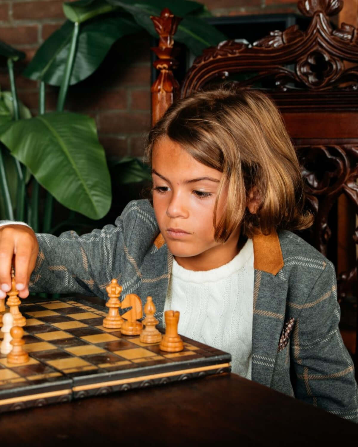 Stylish kid in smoky windowpane blazer, focused on a chess match, exuding confidence and sophistication.