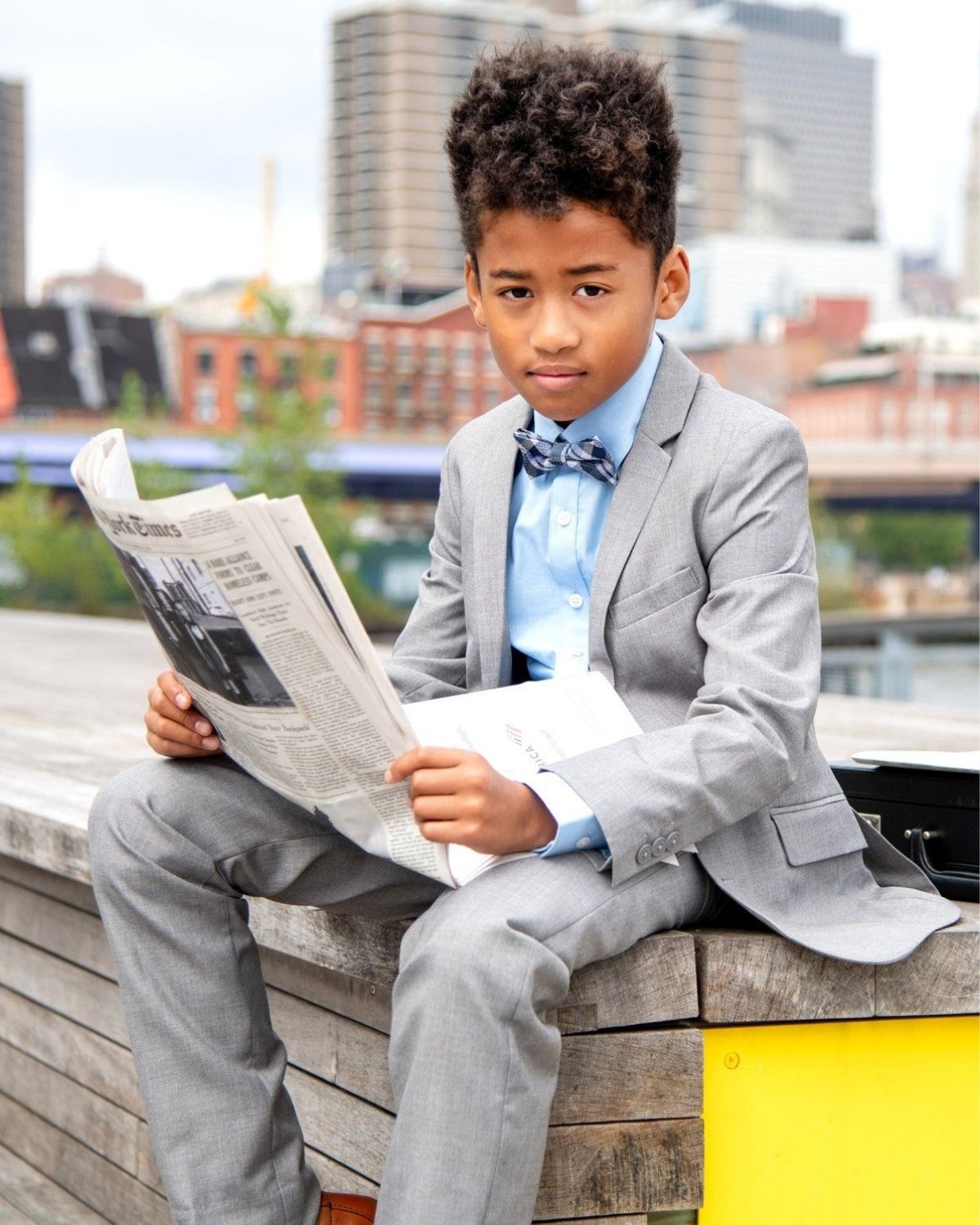 Young boy in a grey suit reading a newspaper outdoors, showcasing the Blue Standard Button Down shirt.
