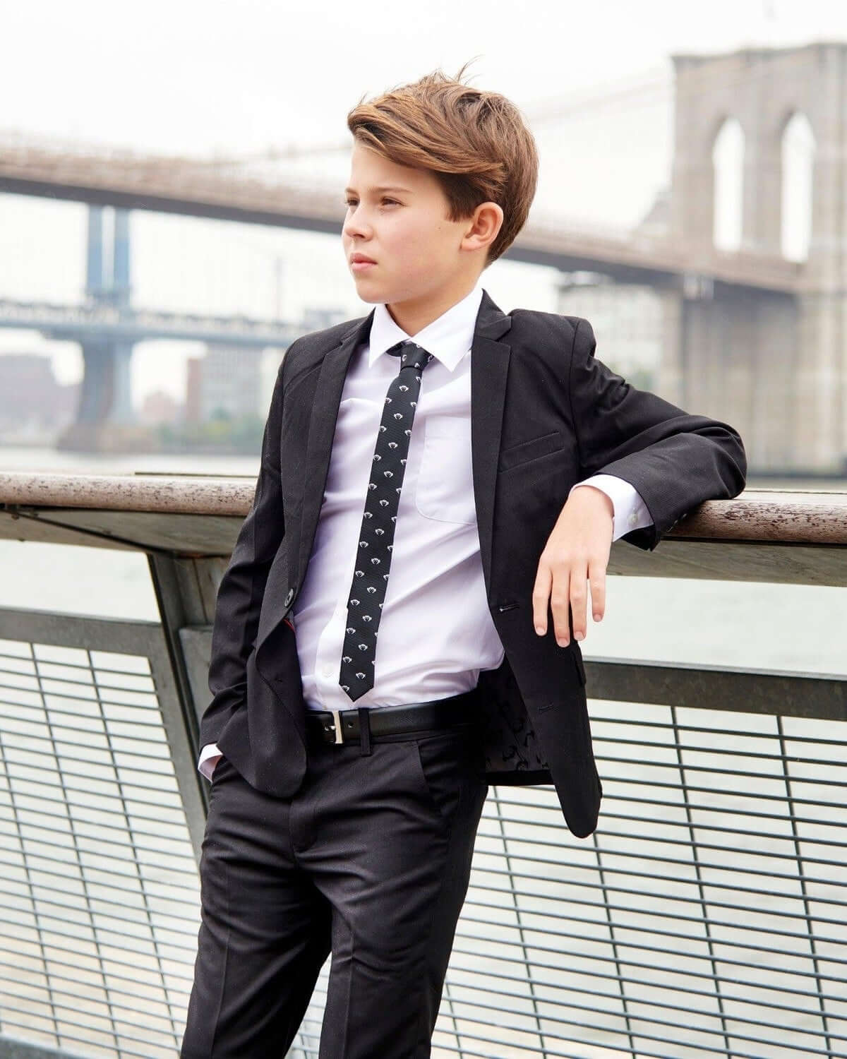A young boy in a suit with a Premium White Standard Button Down shirt, standing by a river with a cityscape in the background.