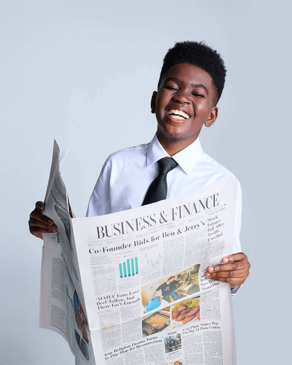 A young boy smiles while holding a newspaper, wearing a Premium White Standard Button Down shirt with a tie.