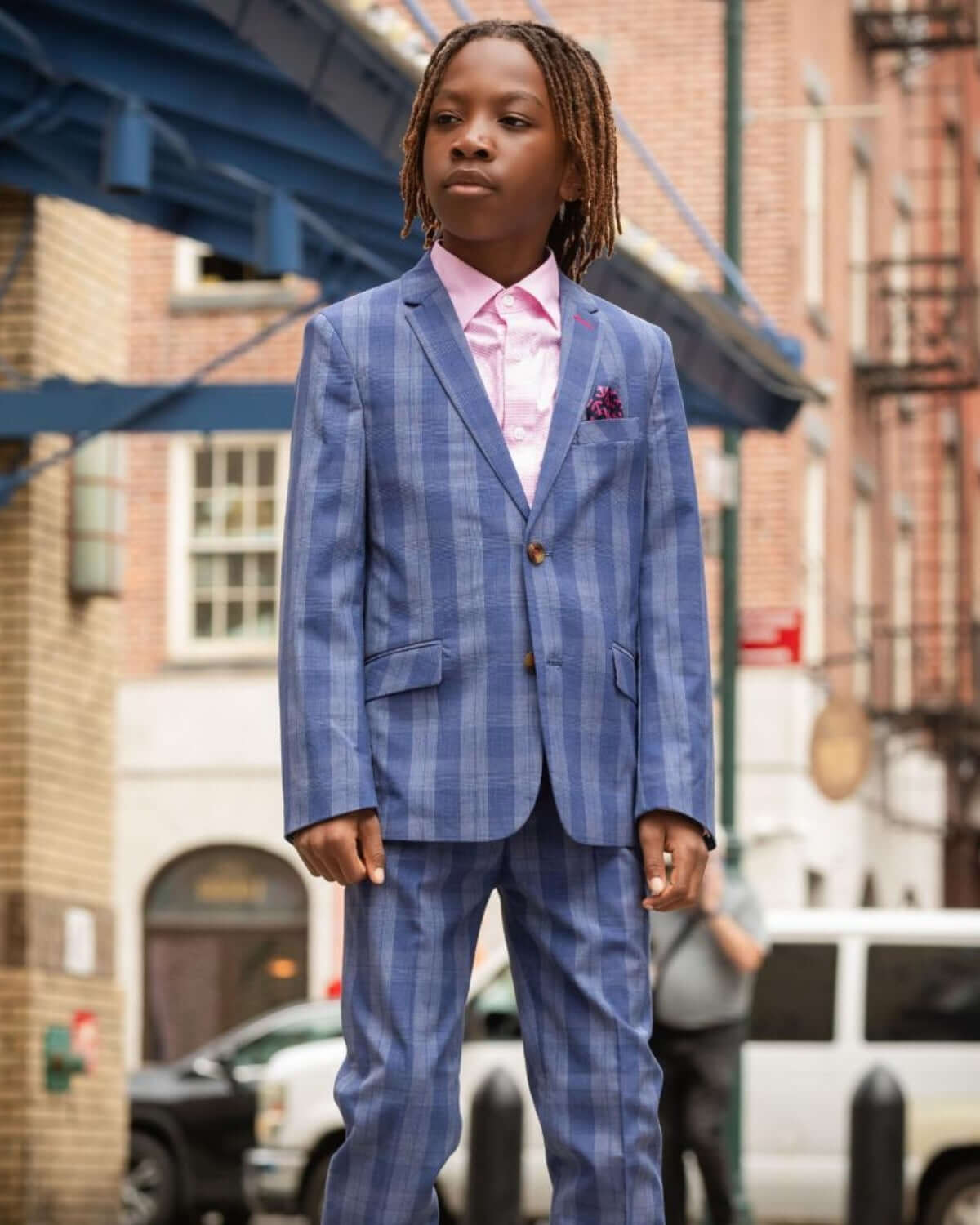 Young boy in Indigo Glen Plaid Stretchy Mod Suit, styled with a pink shirt and confident look on city street.