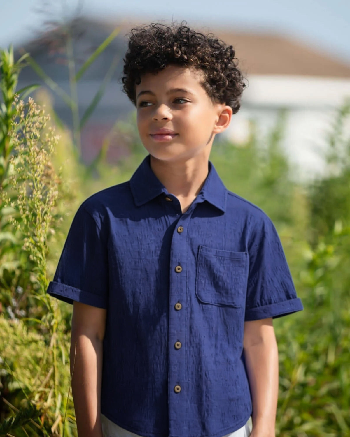 Child wearing a blue shirt standing outdoors with greenery in the background