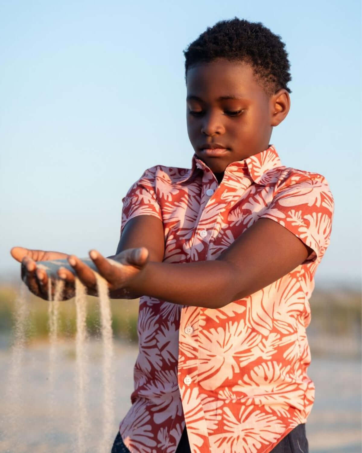 A boy in a vibrant Clay Garden Day Party Shirt, playfully letting sand flow through his hands, enjoying sunny outdoor moments.