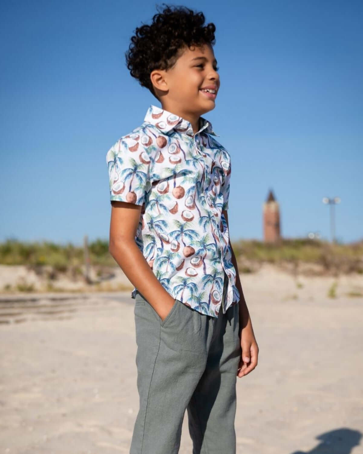 Boy wearing a Coconut Palms Day Party Shirt with a vibrant tropical design on a beach.