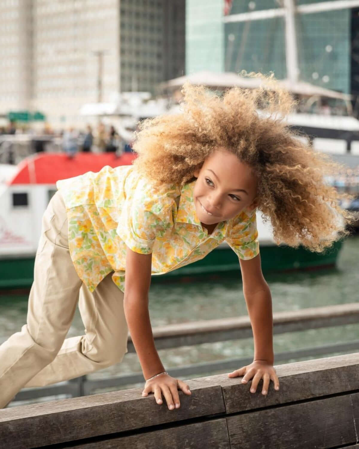 A joyful boy climbing on a railing in a vibrant Spring Bouquet shirt, showcasing style and playfulness by the water.