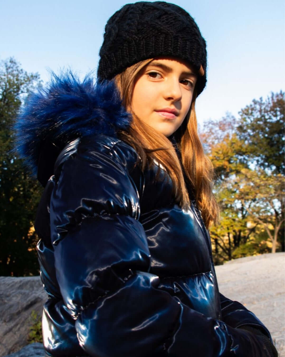 Chic girl in a black cable knit beanie, brightening up a shiny puffer coat while enjoying a crisp fall day.