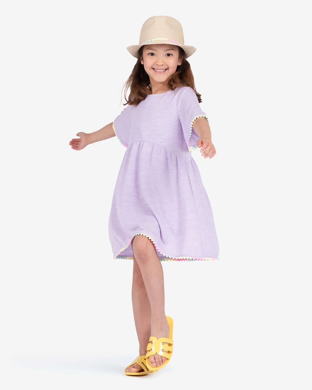 A joyful girl twirls in a lavender dress and stylish fedora, showcasing chic kids clothes with playful flair.
