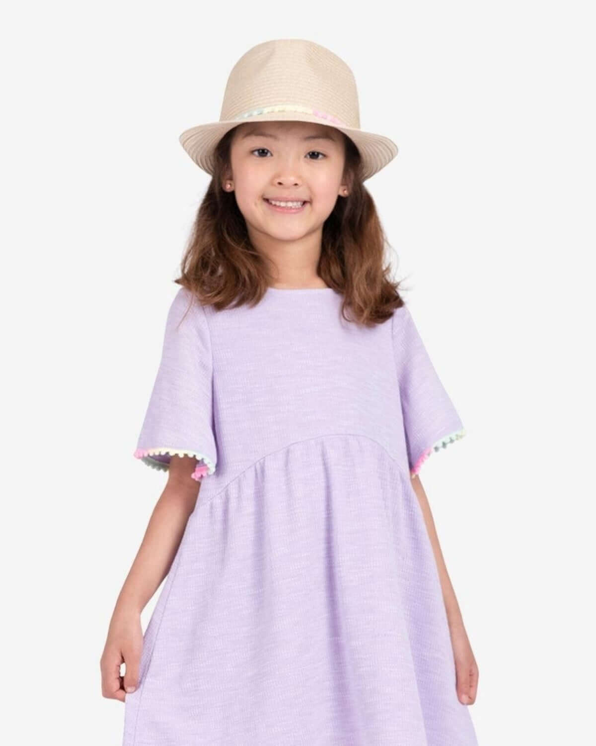 Cute girl in a lilac dress, proudly sporting a stylish natural fedora hat with pink pom-poms for a fun summer look.