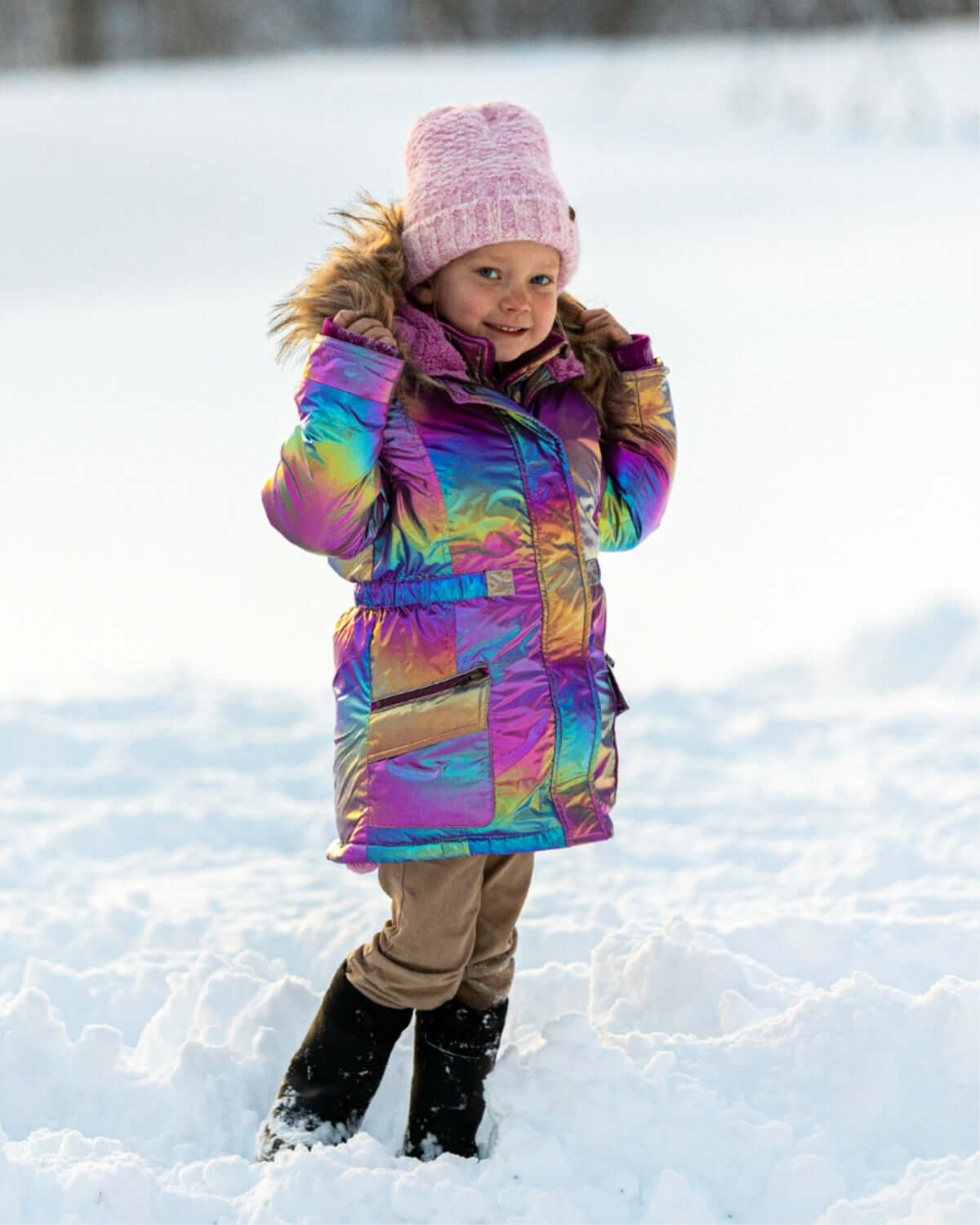 Little girl in vibrant rainbow winter jacket and cozy pink beanie, ready for snow adventures in stylish kids clothes.