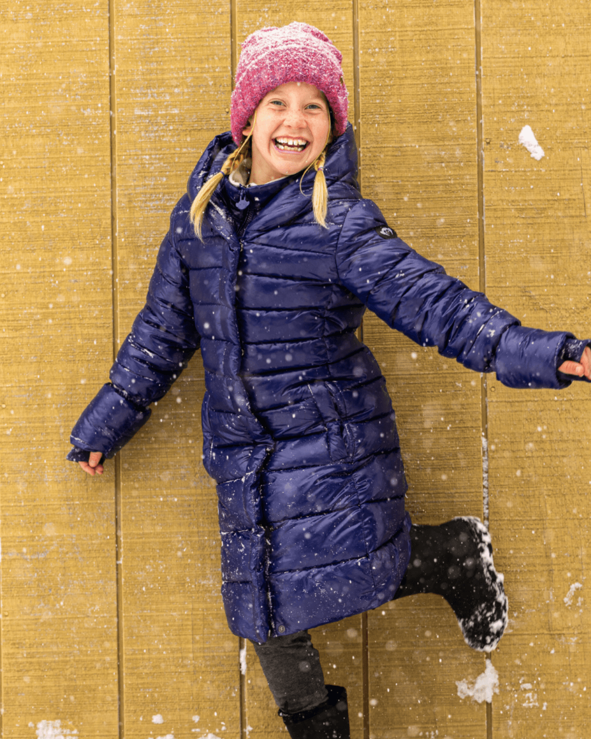 Cheerful girl in a cozy purple puffer coat and pink beanie, enjoying snowy fun and ready for winter adventures.