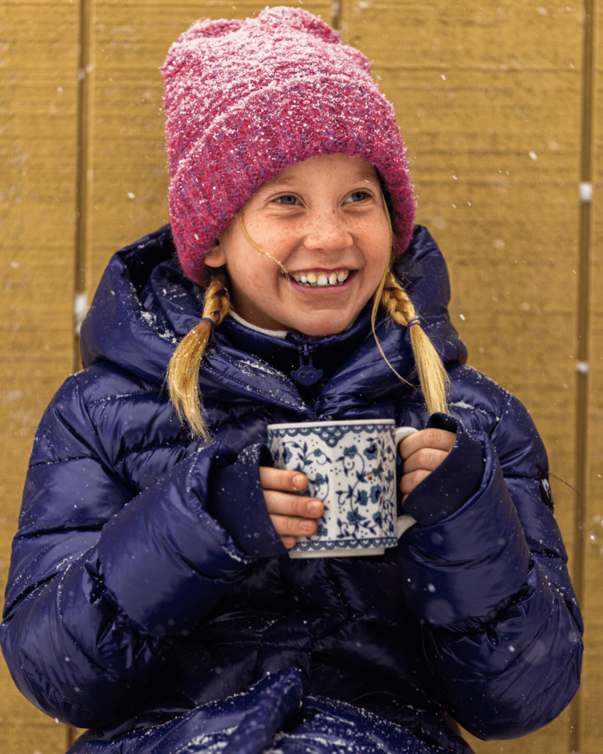 Happy girl in a cozy winter jacket, sipping from a mug while wearing a vibrant pink beanie, perfect for stylish kids' warmth.