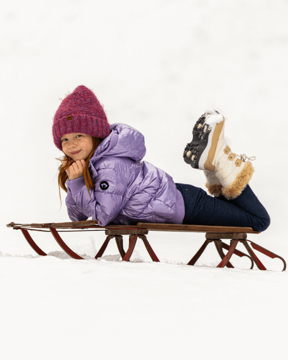 Charming girl in a cozy wine red Tracy beanie enjoying a snowy sled ride, showcasing stylish kids clothes for winter fun!