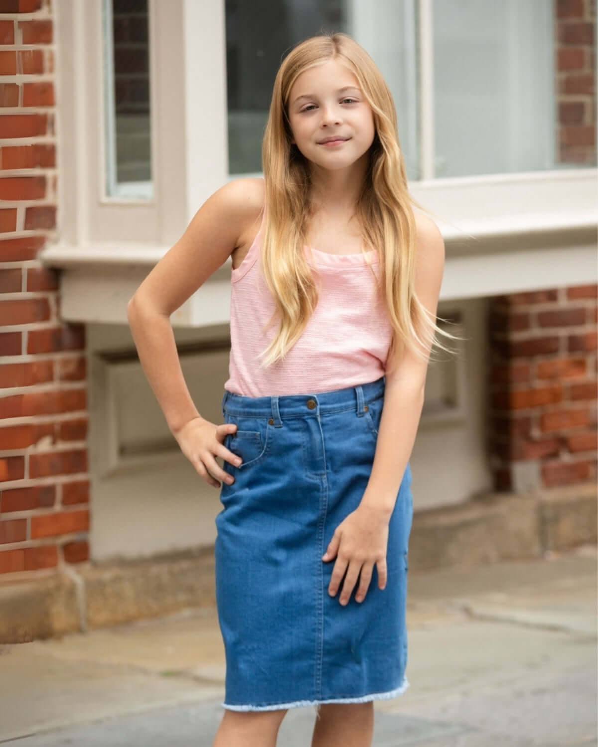 Girl wearing Blue Jean Rhodes Midi Denim Skirt, styled with a pink tank top, against a brick wall background.
