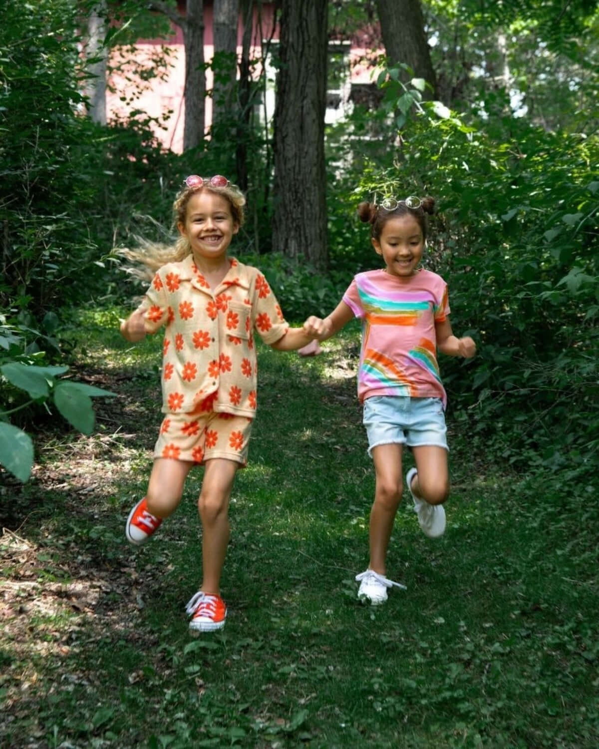 Two joyful girls in colorful kids clothes run hand-in-hand through a sunny forest path, embracing summer fun.
