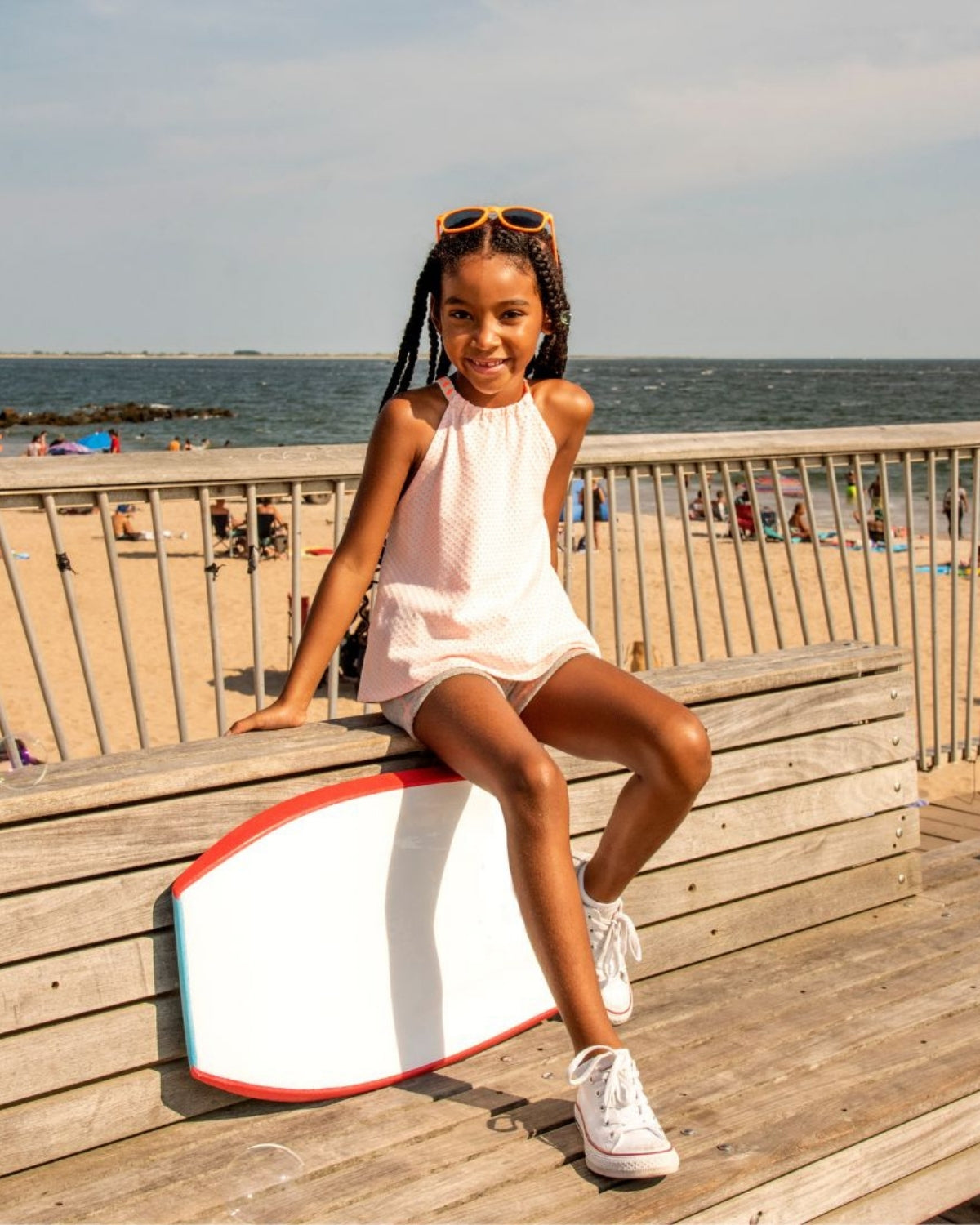 A cool girl in stylish summer clothes poses on a beach bench, ready for fun in the sun with a surfboard nearby.