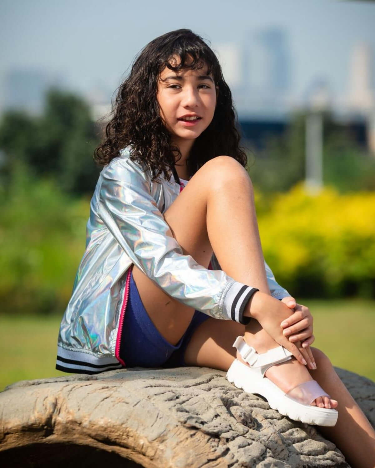 Chic girl in shiny bomber jacket and sandals, rocking playful summer vibes in a sunny park. Perfect kids' clothes for style and comfort!