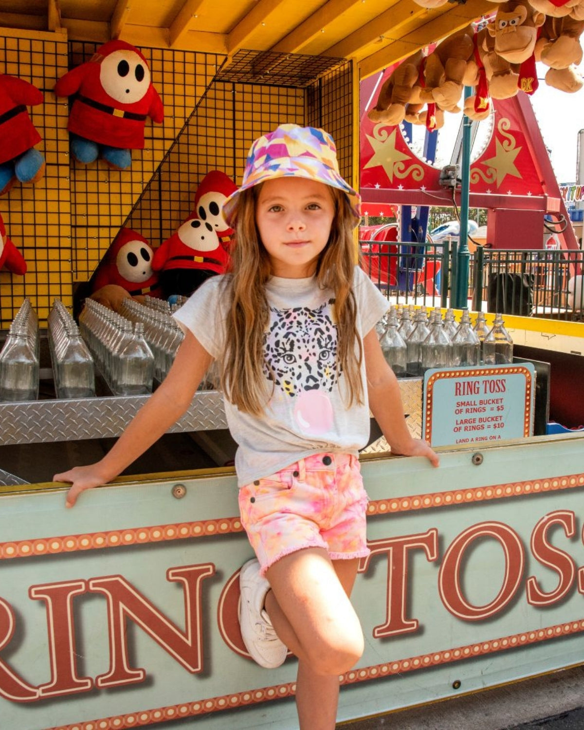 Stylish girl in neon shorts and a colorful bucket hat, enjoying summer fun at a carnival—perfect kids clothes for adventurous spirits!