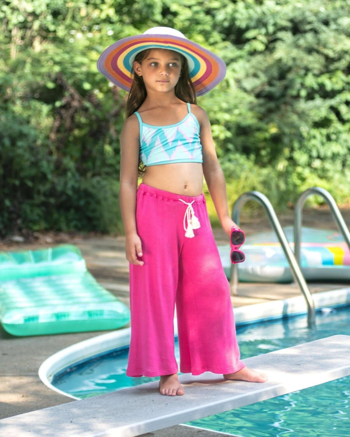 Girl wearing Radiant Pink Beach Pant and a colorful hat by the pool, enjoying summer vibes.