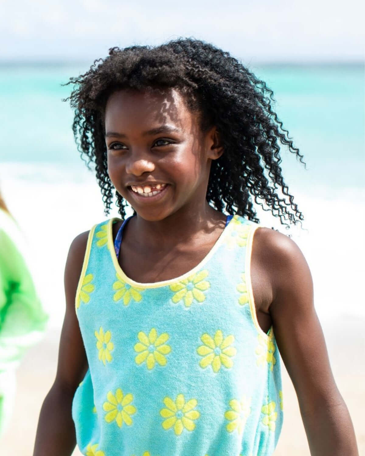 A girl wearing the Blue Daisies Mia Dress with yellow daisies, smiling joyfully at the beach.
