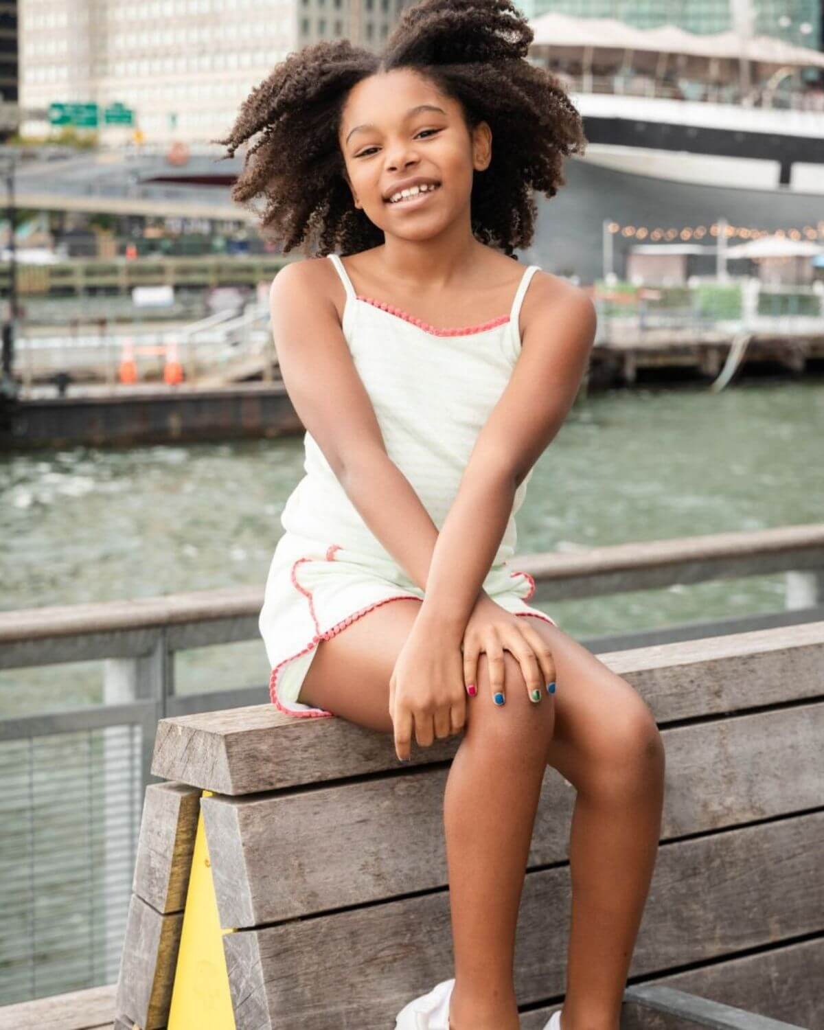 Light Green Stripe Novia Romper worn by a smiling girl sitting on a wooden bench near water.