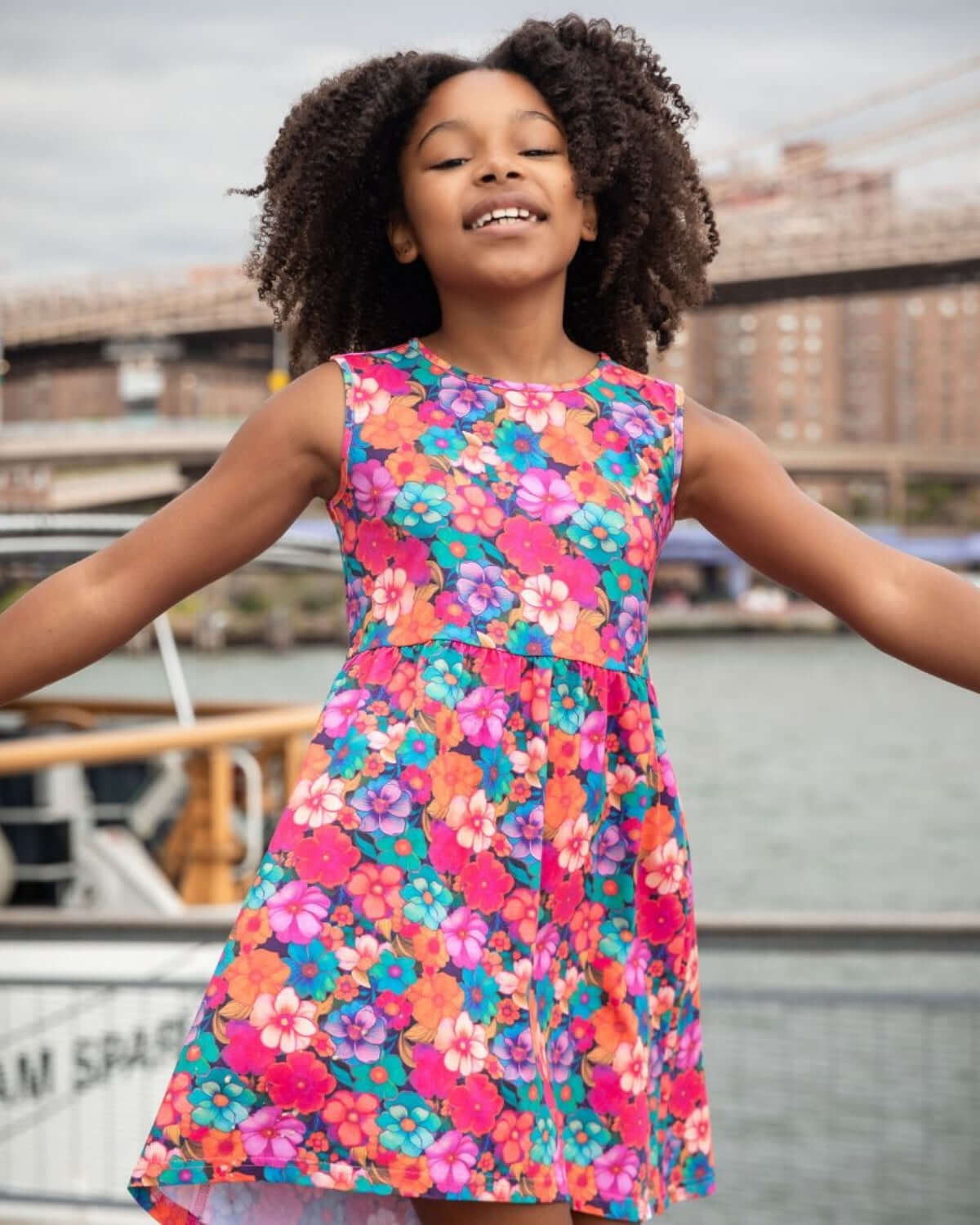 Girl wearing a colorful dress by the water, showcasing the Summer Bloom Naxios Dress with vibrant floral design.